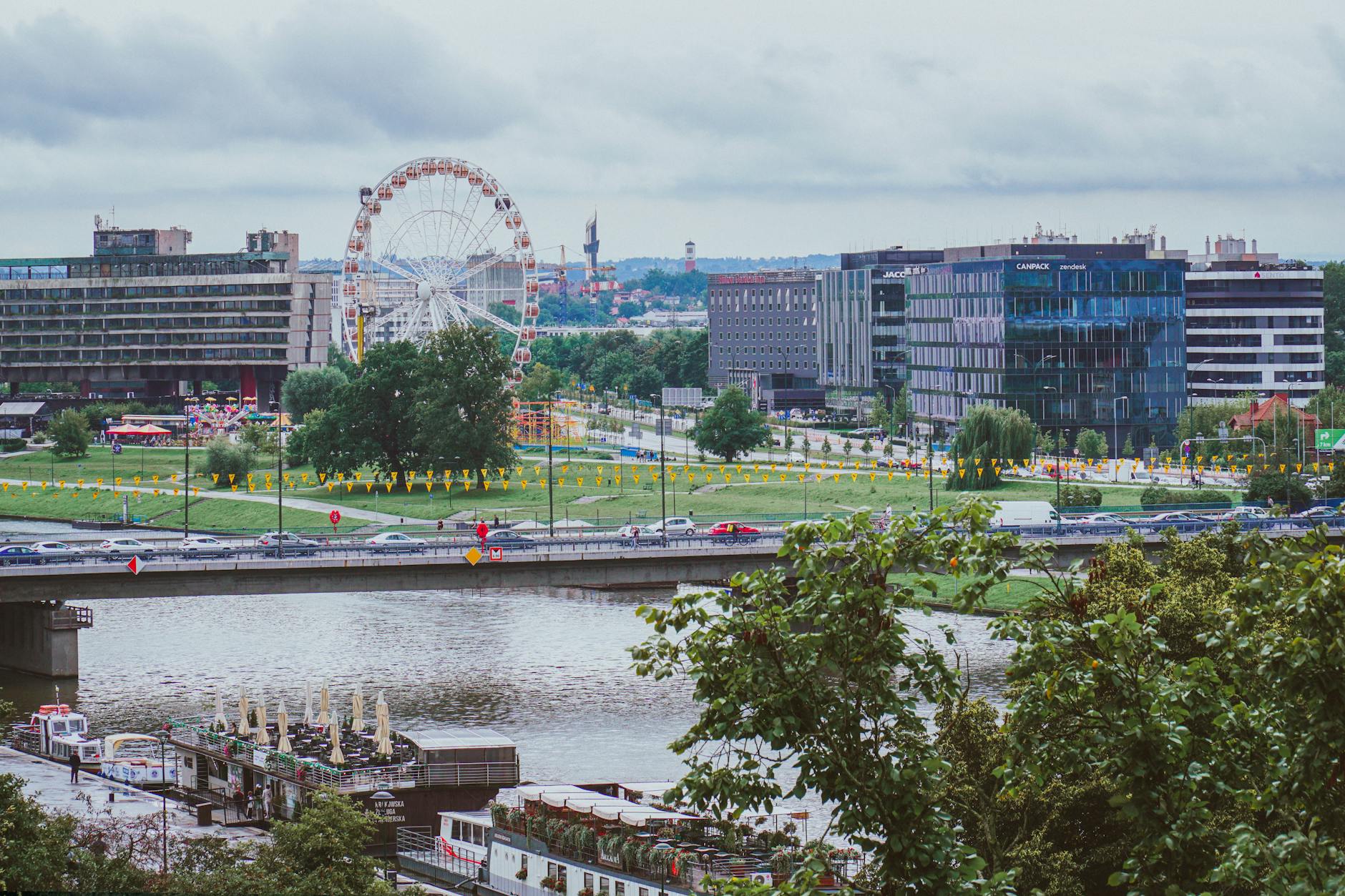 A vibrant cityscape of Kraków featuring a ferris wheel and river under cloudy skies.