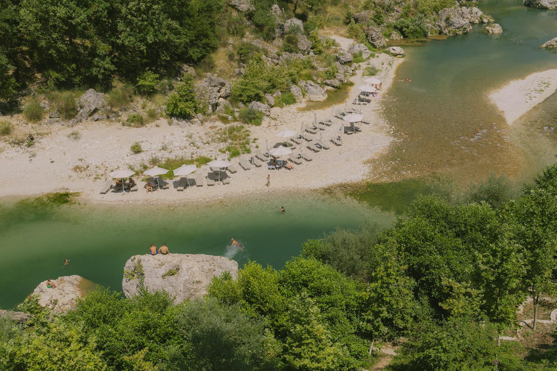 Peaceful riverside scene with sun umbrellas and lush greenery in Tepelenë, Albania.